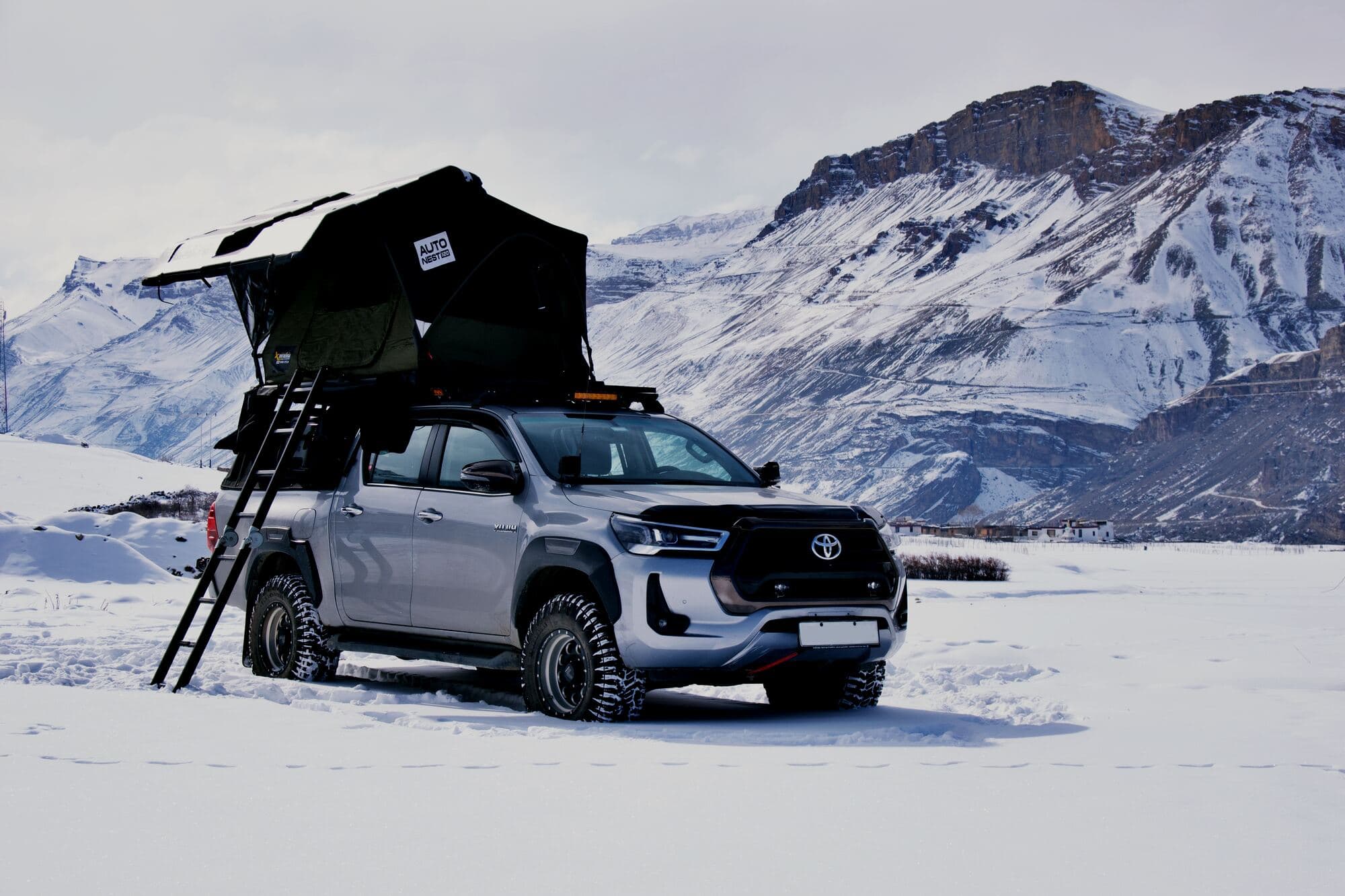 A Toyota Hilux with an open hardshell rooftop tent on a frozen Himalayan plain beneath snow peaks